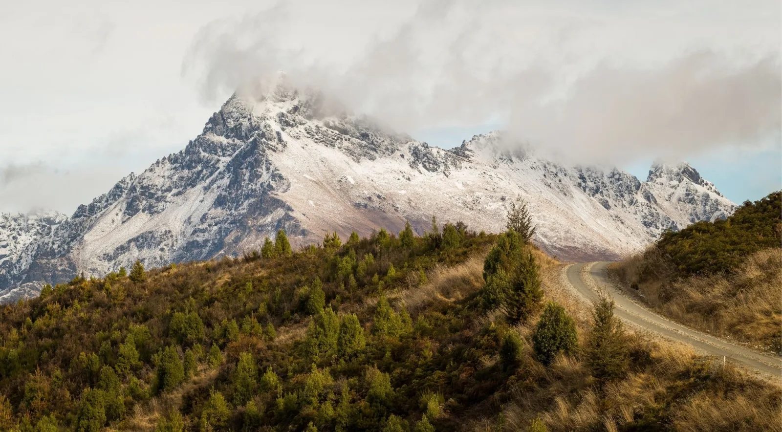 Cloud tearing across a snow-dusted summit above Glenorchy, with beech scrub and a farm road below