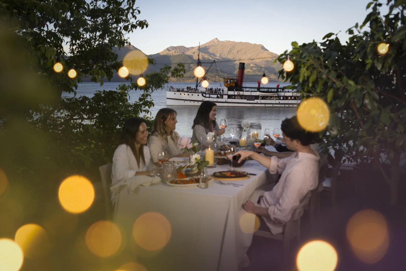 Guests dining under festoon lights on the Colonel's Homestead terrace as the TSS Earnslaw lies at the Walter Peak jetty behind them