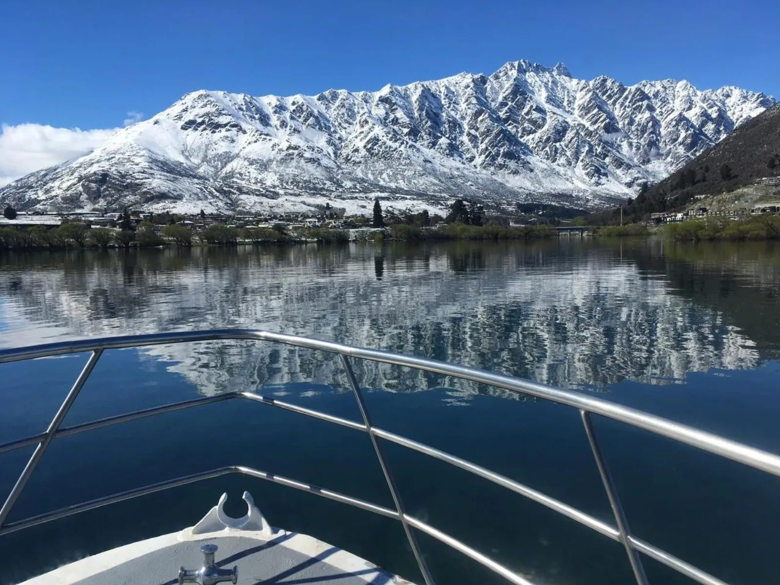 The Remarkables under fresh snow reflected in a mirror-calm Lake Whakatipu, seen over the boat's bow rail