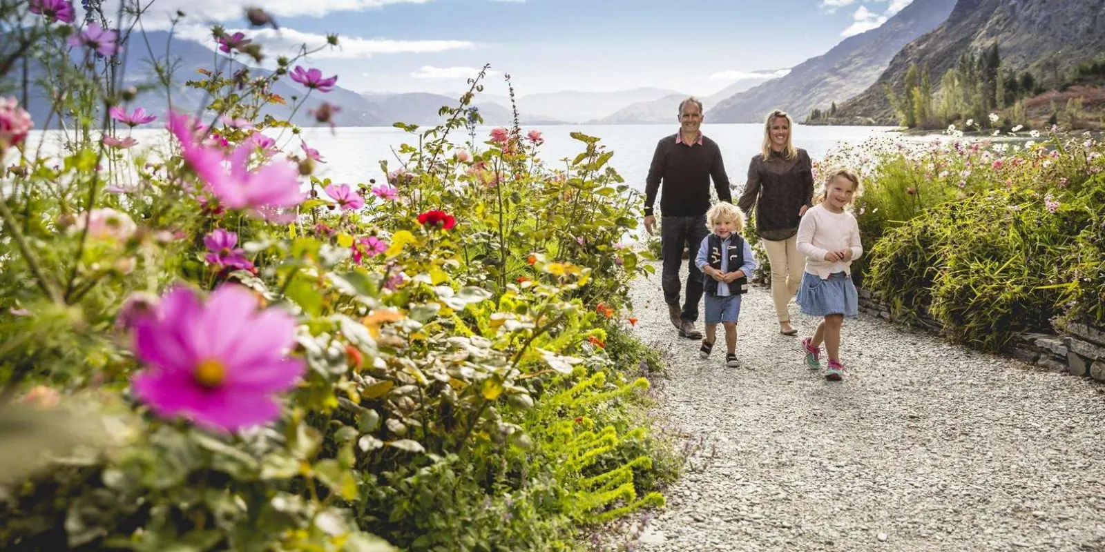 A family walking a gravel path through the Walter Peak flower gardens, cosmos in the foreground and Lake Whakatipu beyond