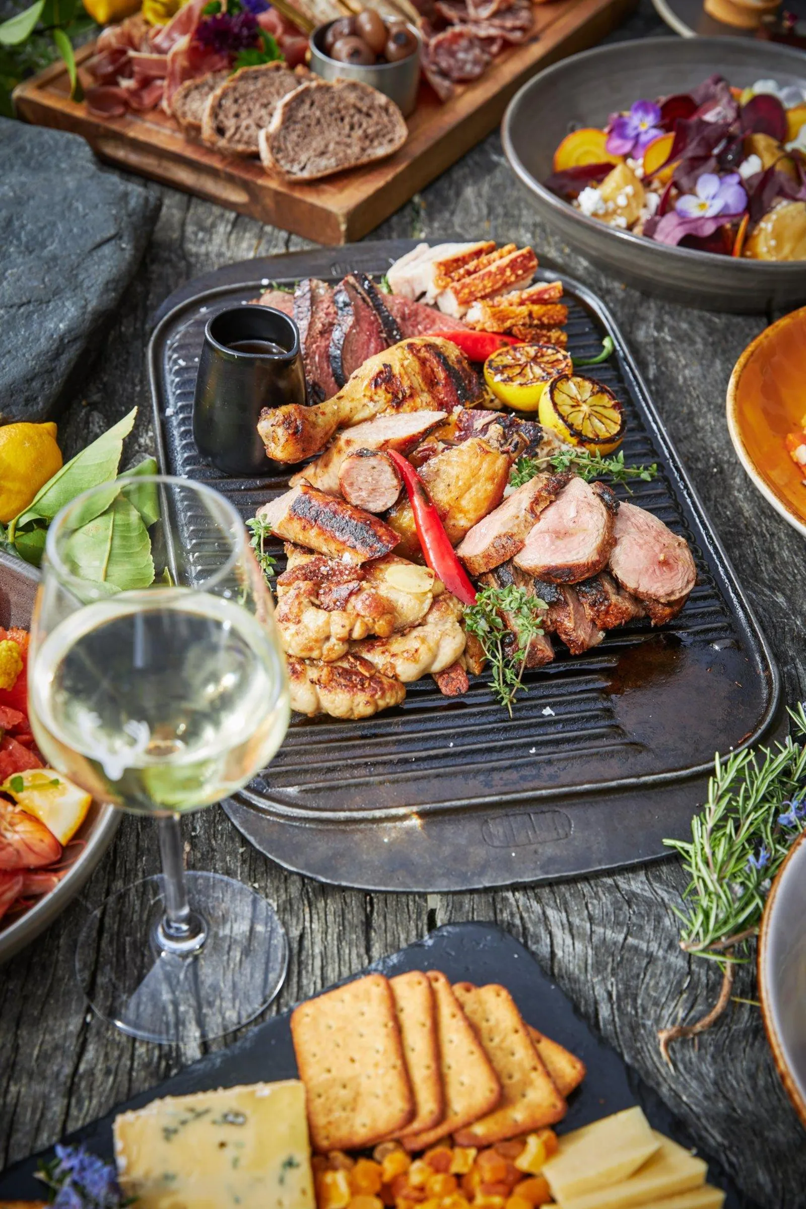 Grilled meats and vegetables resting on a griddle plate beside salads and a glass of white wine at Walter Peak