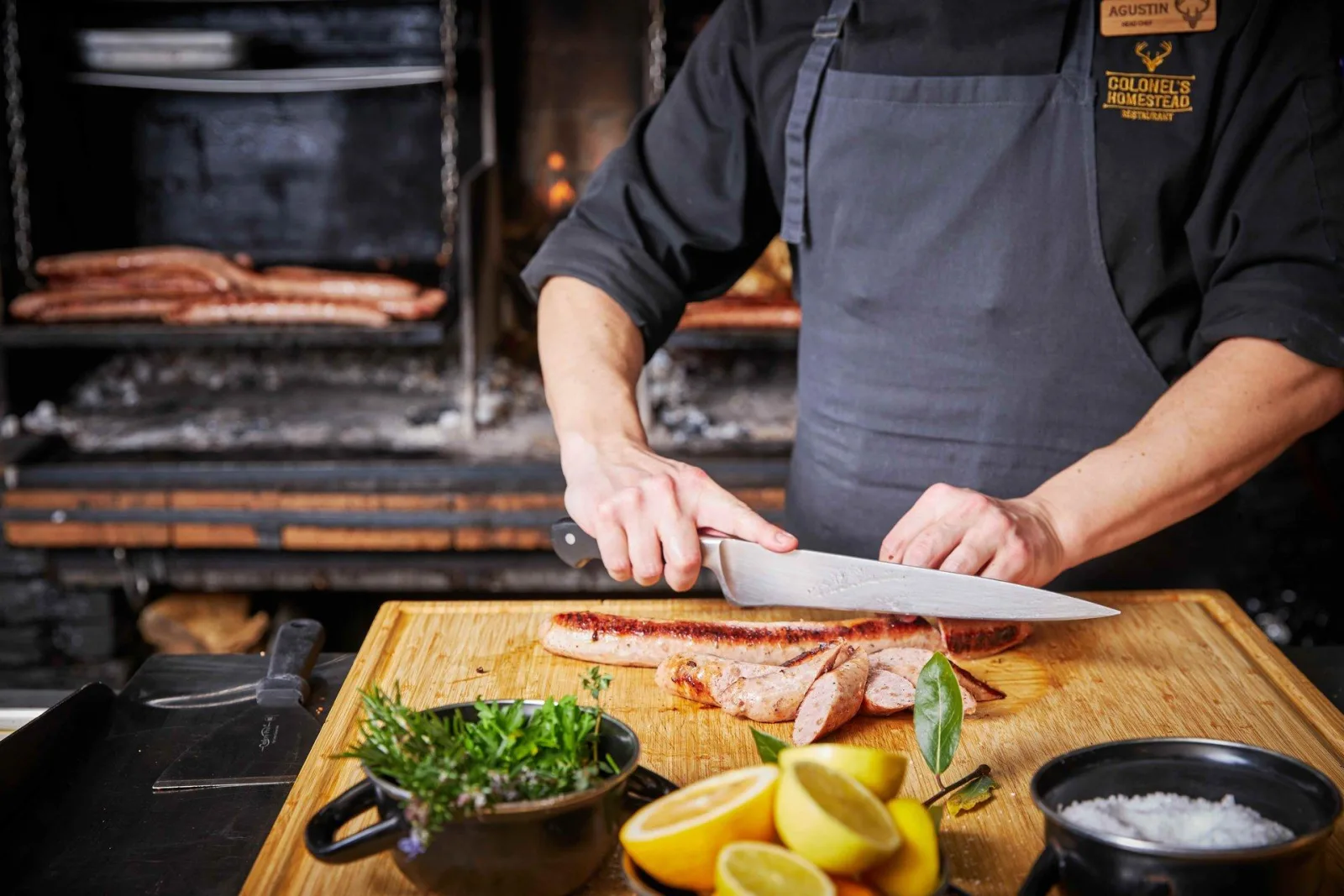 A chef in a Colonel's Homestead apron slicing rare beef beside the open wood-fired grill